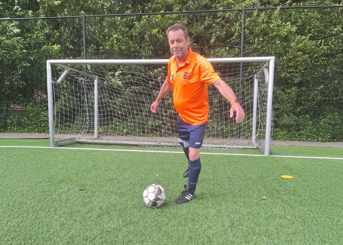 Een man in een oranje shirt en een blauwe korte broek die tegen een voetbal trapt