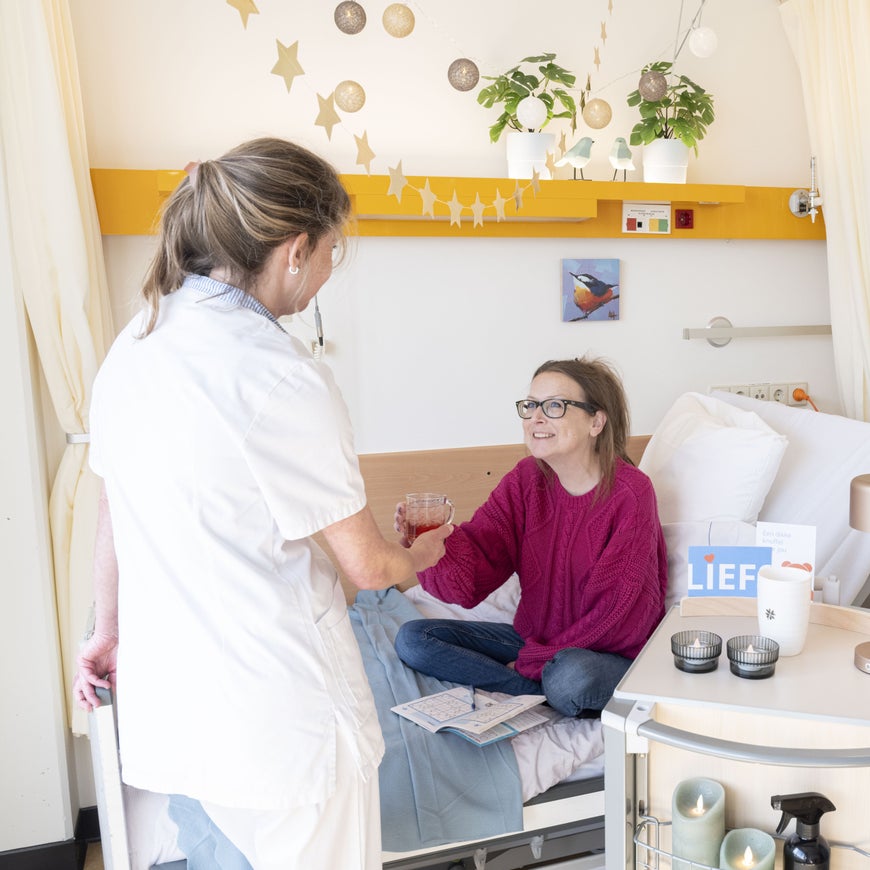 Een vrouw in een ziekenhuisbed krijgt een glas drinken aangereikt van een zorgverlener.