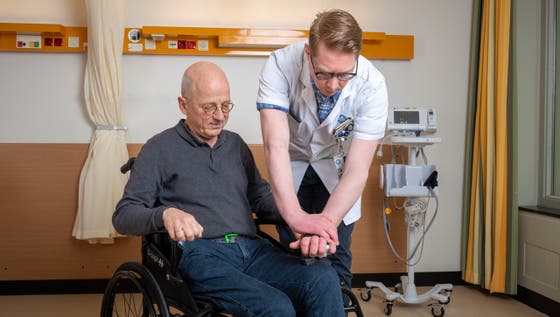 Een man in een rolstoel, terwijl een arts zijn hand vasthoudt