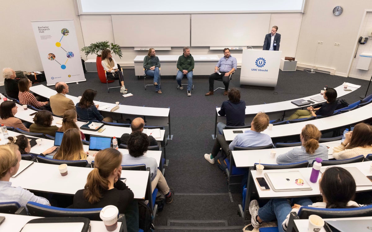 Studenten zitten in een college zaal en kijken naar een groepje mensen op stoelen die in gesprek zijn.