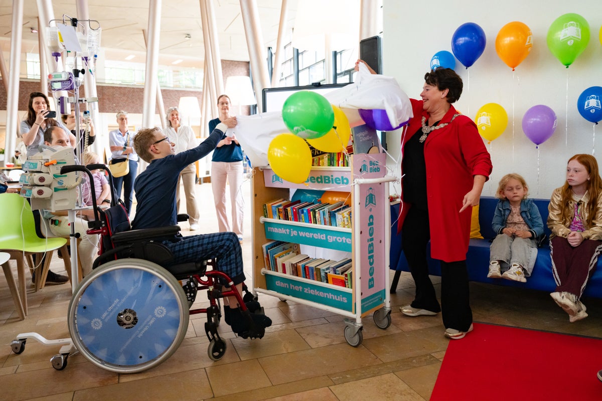Een jonge patiënt in een rolstoel en Sharon Dijksma, burgemeester van Utrecht, onthullen samen de BedBieb, een rijdende bibliotheek voor kinderen in het WKZ die bedgebonden zijn. 