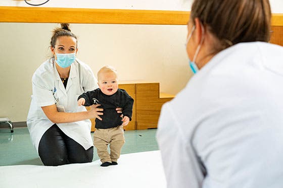 Een kinderarts begeleidt een jong kind dat leert lopen tijdens een medische afspraak, beiden dragen mondkapjes in een klinische omgeving