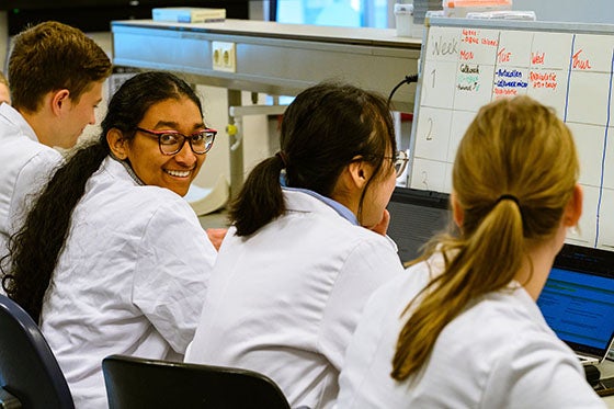 Studenten in witte labjassen werken aan laptops en whiteboards in een klaslokaal. Een studente met bril glimlacht naar de camera