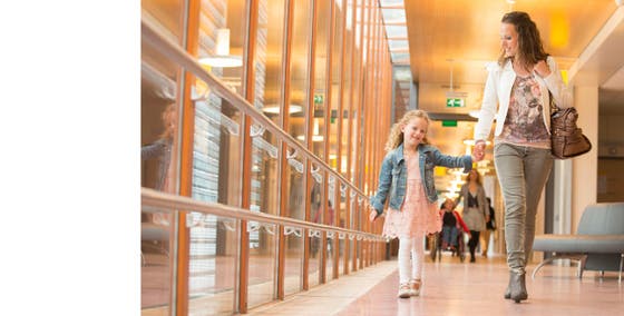 Een meisje in een roze jurk en spijkerjasje loopt met een vrouw door een gang van het UMC Utrecht